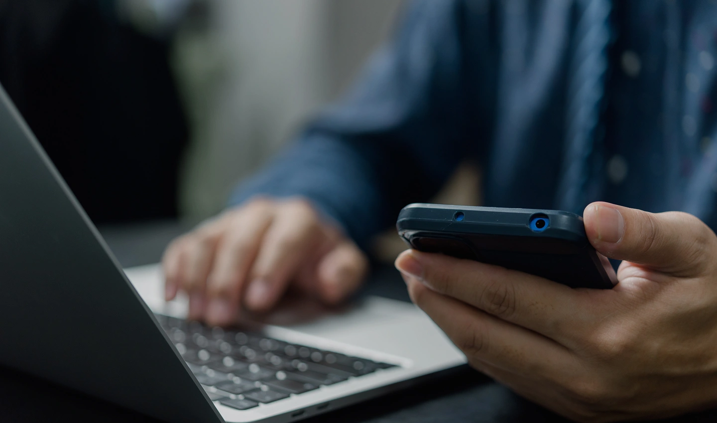 Person typing on a laptop while holding a smartphone near the keyboard, multitasking setup.