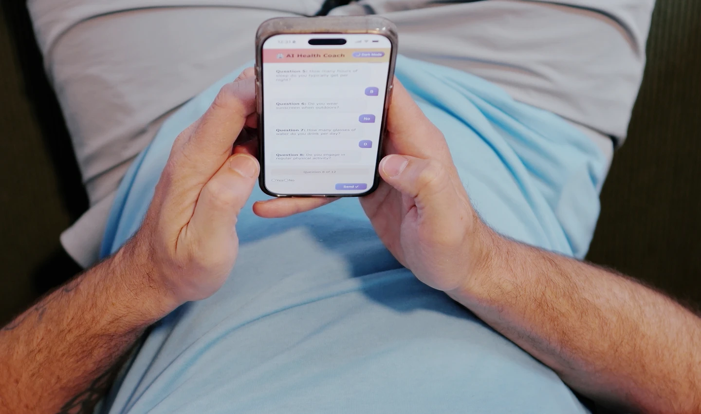 Person seated, holding a smartphone showing an AI Health Coach chat interface with several questions on screen.