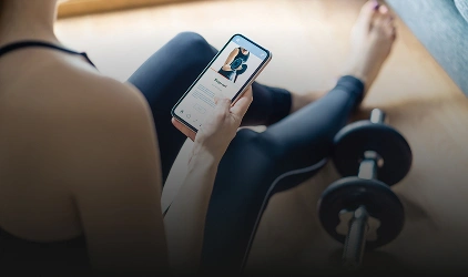 Person wearing athletic clothing looks at a fitness app on a phone, with a dumbbell nearby during a workout session in a gym area.