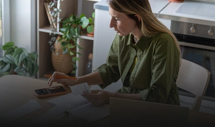 Woman in a green shirt sits at a kitchen table, reviewing papers with a smartphone in hand.