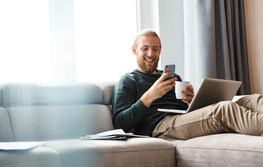 Bearded man on a sofa at home, scrolling on his phone with a mug in hand and a laptop nearby.