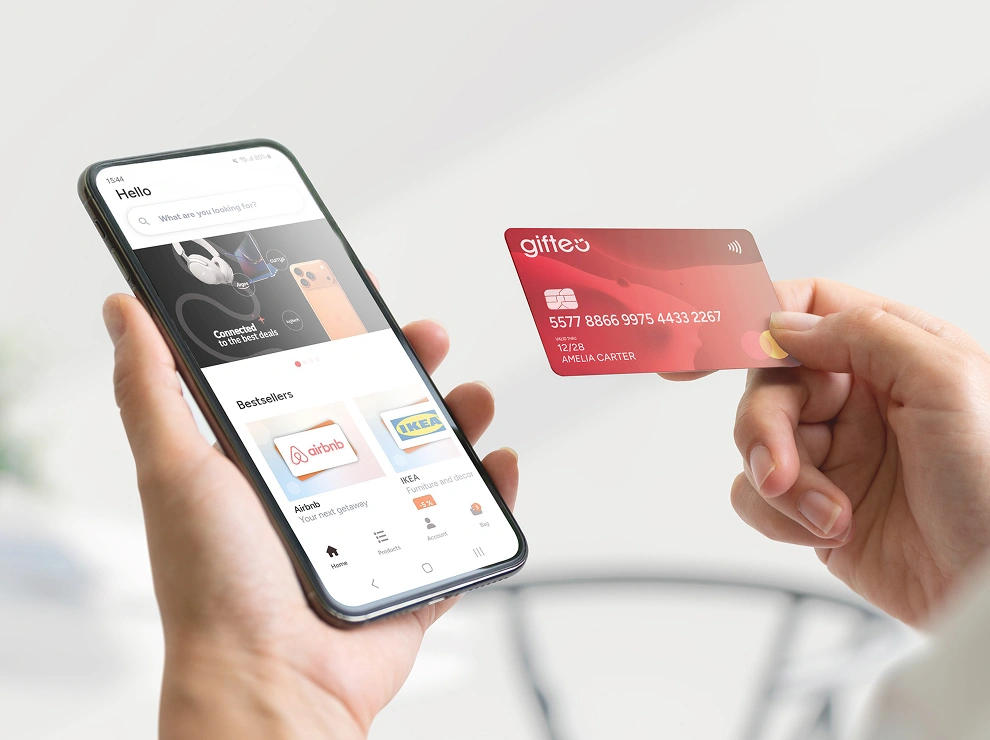 Person holds a smartphone displaying a shopping app while another hand holds a red gift card to pay.